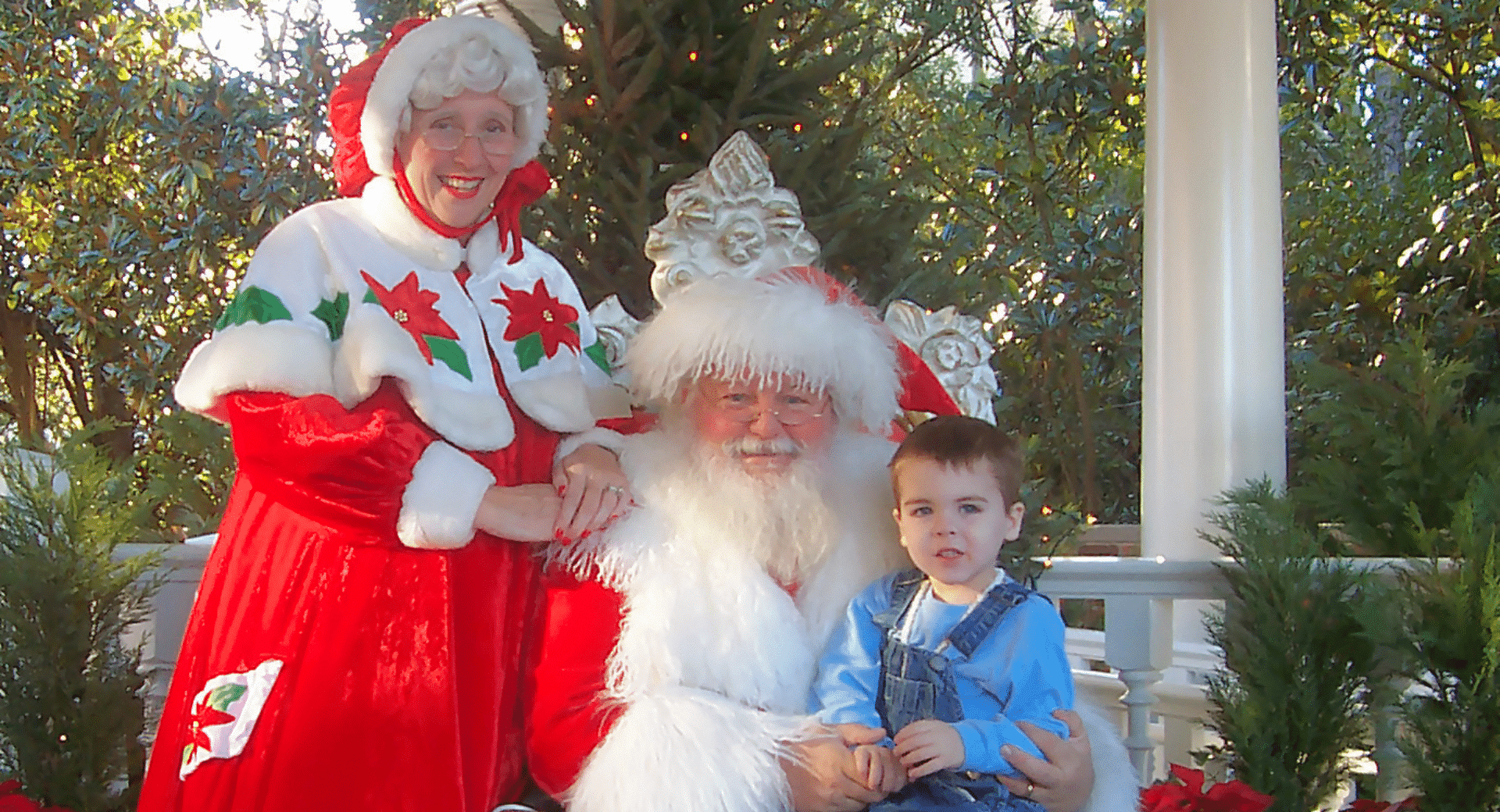 A young boy in denim overalls sits on Santa Claus’s lap in a festive outdoor setting at Epcot. Santa and Mrs. Claus are both dressed in traditional red and white holiday attire with poinsettia accents, posing together in a decorated gazebo surrounded by greenery.