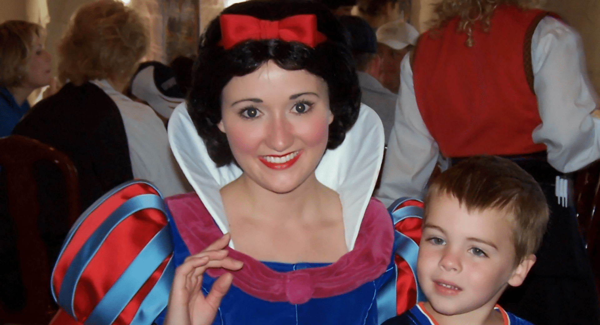 A young boy with short brown hair stands next to Snow White during a character meal. Snow White is wearing her classic blue, red, and yellow gown with a high white collar and a red bow in her hair, smiling warmly at the camera while the boy looks on with a soft expression.