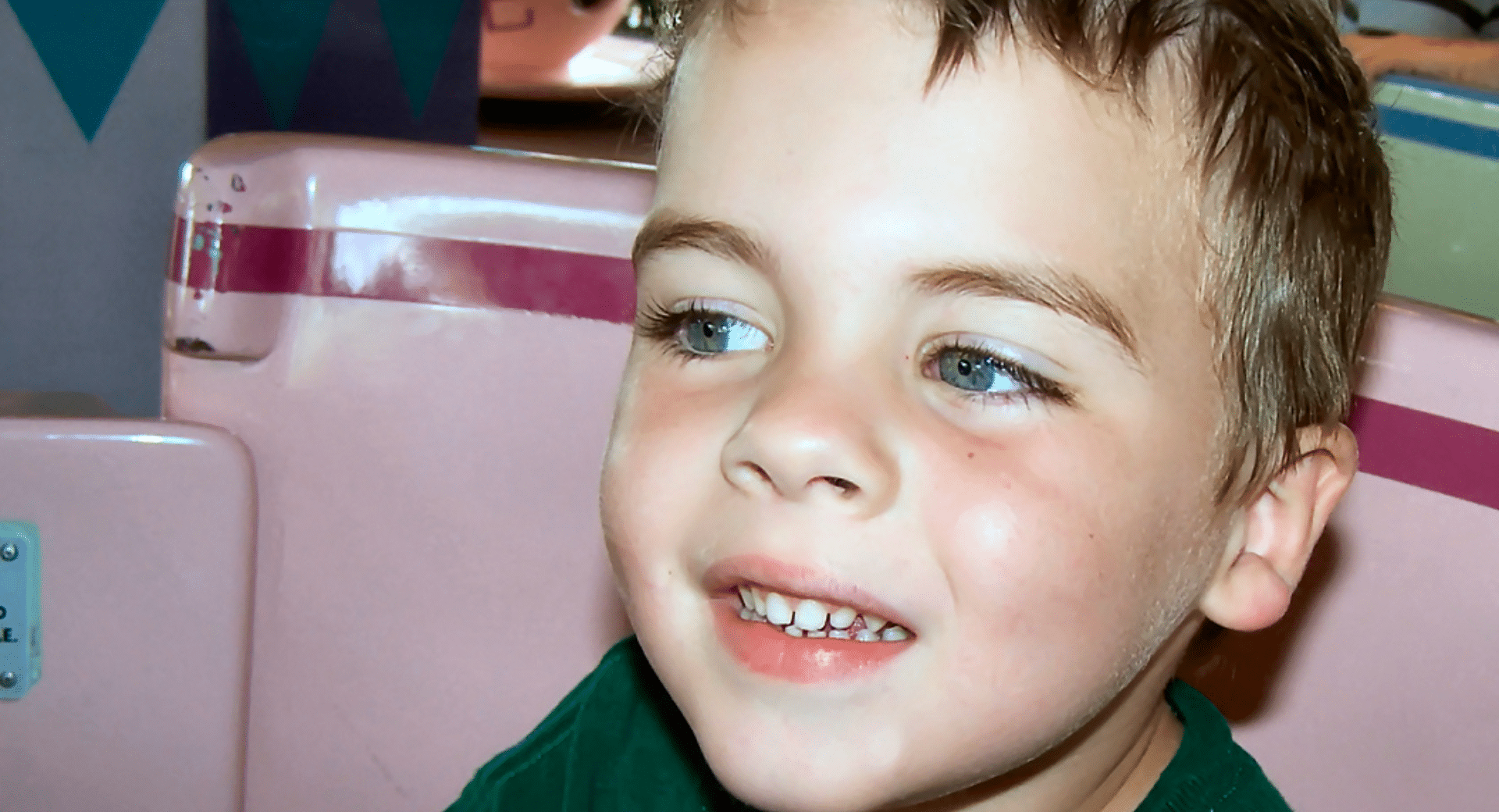 A close-up, candid photo of a young boy with light brown hair and blue eyes, smiling brightly while riding the Mad Tea Party attraction. He is sitting in a pink teacup, and the background shows the whimsical, colorful patterns of the ride.